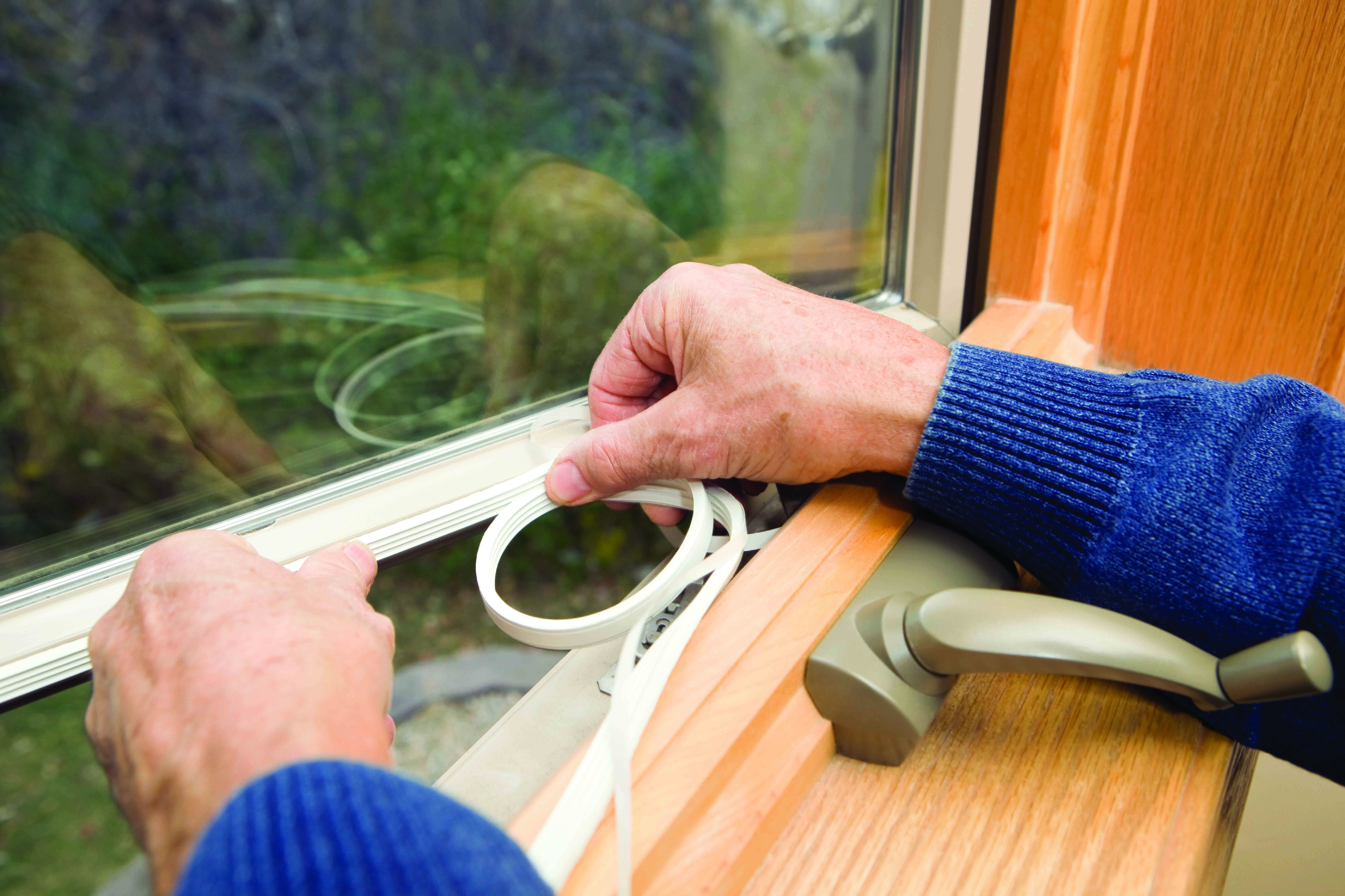 An older white person in a blue sweater adds weatherstripping to an open window to weatherize their home.